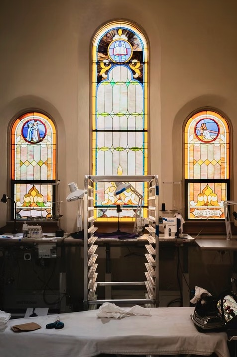 Work tables and garment racks beneath three tall stained glass windows in the former church sanctuary